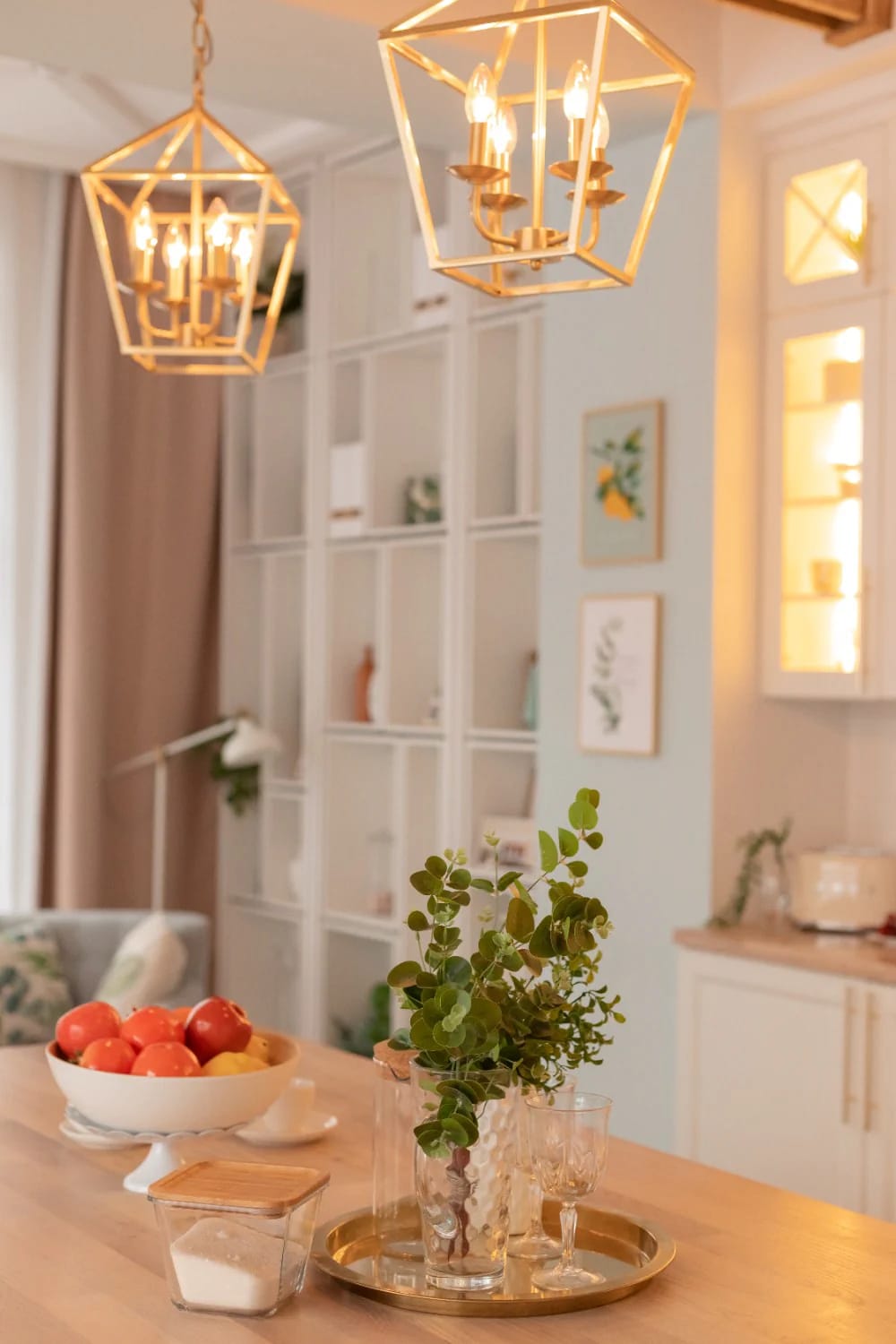 Kitchen Remodel Inspiration With Layered Lighting Kitchen remodel inspiration showing two geometric pendant lights above a wood dining table, with a fruit bowl, plant arrangement, and illuminated cabinetry in the background