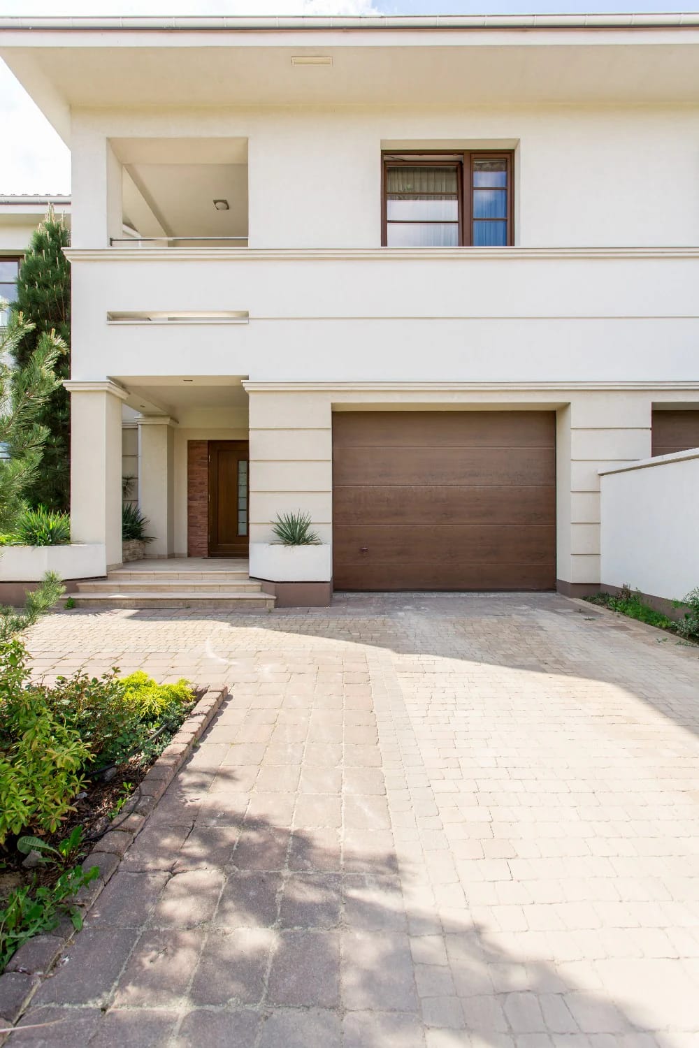 Two-story narrow-lot house front with a prominent brown garage door, recessed entry, upper balcony, window, and paved driveway illustrating how the garage sets the front elevation