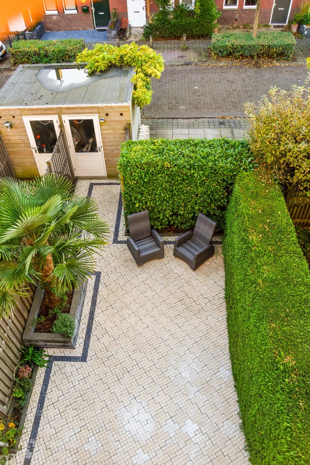 Paved courtyard garden with two chairs, raised planters, tall hedges, tropical plants, and a small wooden garden building, illustrating outdoor living in narrow lot house plans