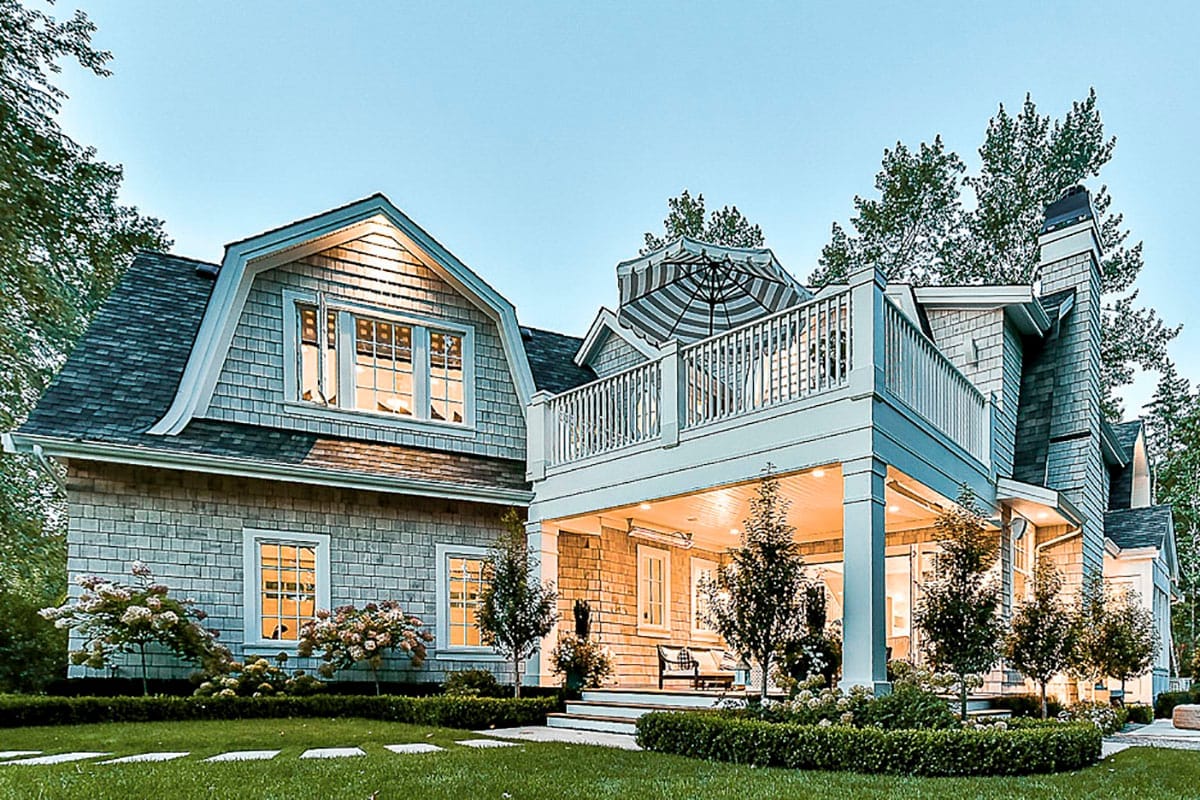 Shingle-style home with gambrel roof, upper deck, and covered porch surrounded by lush landscaping.