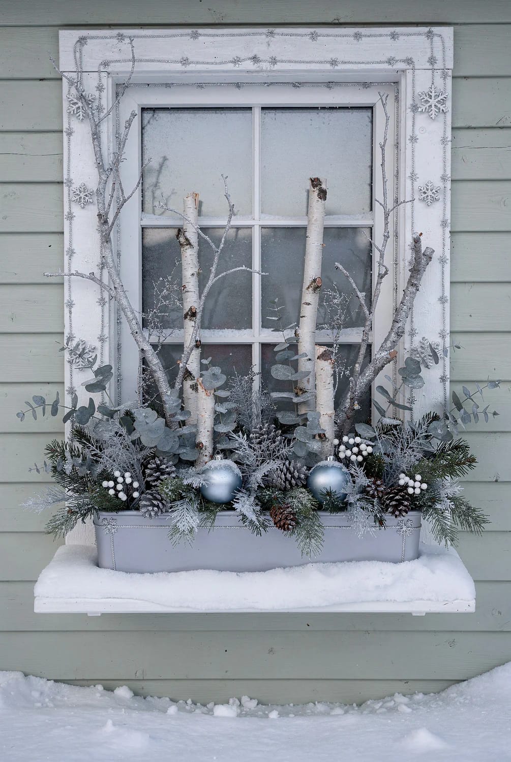 Exterior window box filled with evergreen branches, birch logs, frosted foliage, pinecones, white berries, and silver-blue ornaments beneath a snow-covered sill