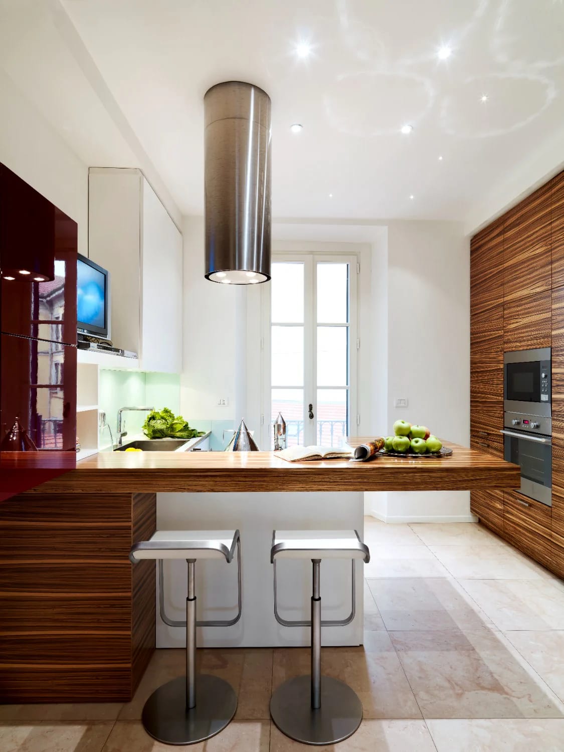 Small Apartment Kitchen With Counter Seating Small apartment kitchen arranged along the walls with wood-grain cabinetry, a long counter, two bar stools, a cylindrical range hood, built-in appliances, a window, and food on the countertop