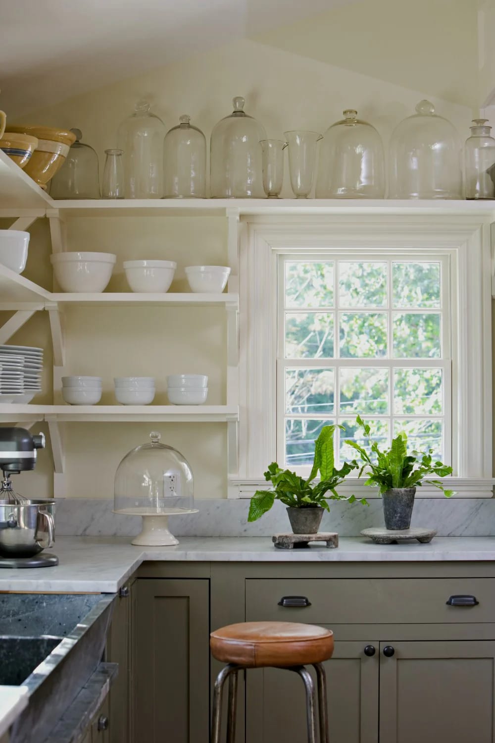 11 Small Apartment Kitchen Ideas That Give You Back Your Counter 2 Small apartment kitchen featuring open shelves filled with glass jars and white bowls, a marble countertop, potted plants by the window, and olive-green lower cabinets.