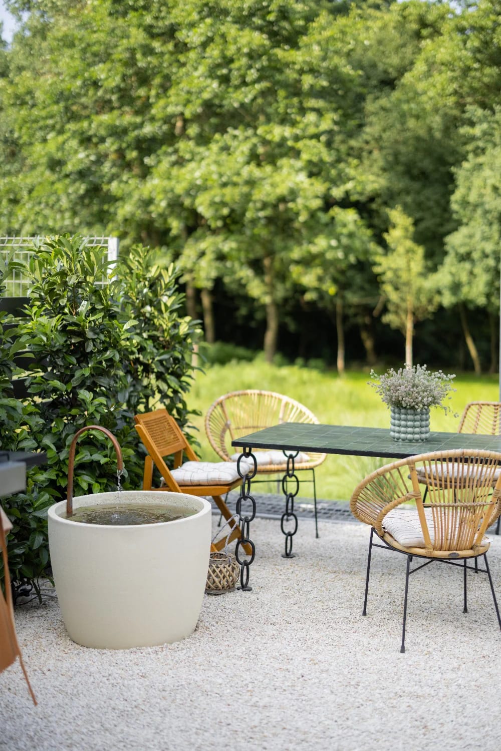 Small Backyard Landscaping With Continuous Gravel Floor Small backyard landscaping idea showing a continuous light gravel floor with wicker chairs, a dark table, potted plants, and a water fountain surrounded by trees