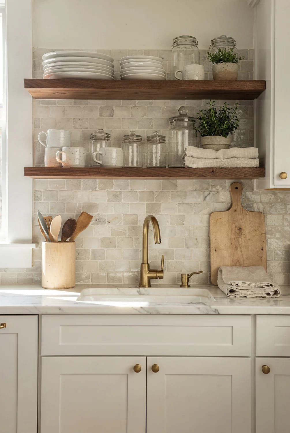 Timeless Kitchen Remodel Open Shelving Kitchen remodel inspiration showing two short walnut open shelves above a sink, styled with stacked white plates, mugs, glass jars, plants, and folded towels against a light tiled backsplash