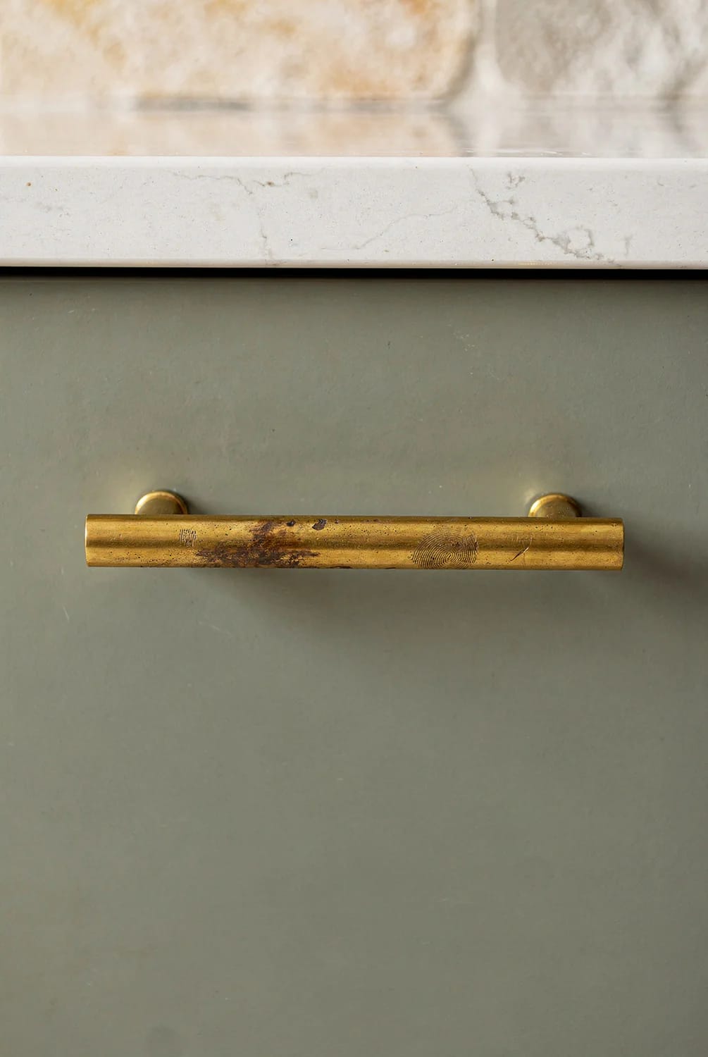 Kitchen Remodel Inspiration That Holds Up After the Trend Passes 3 Close-up of a tarnished unlacquered brass bar pull mounted on a muted green kitchen cabinet beneath a light stone countertop, illustrating a lasting kitchen remodel finish