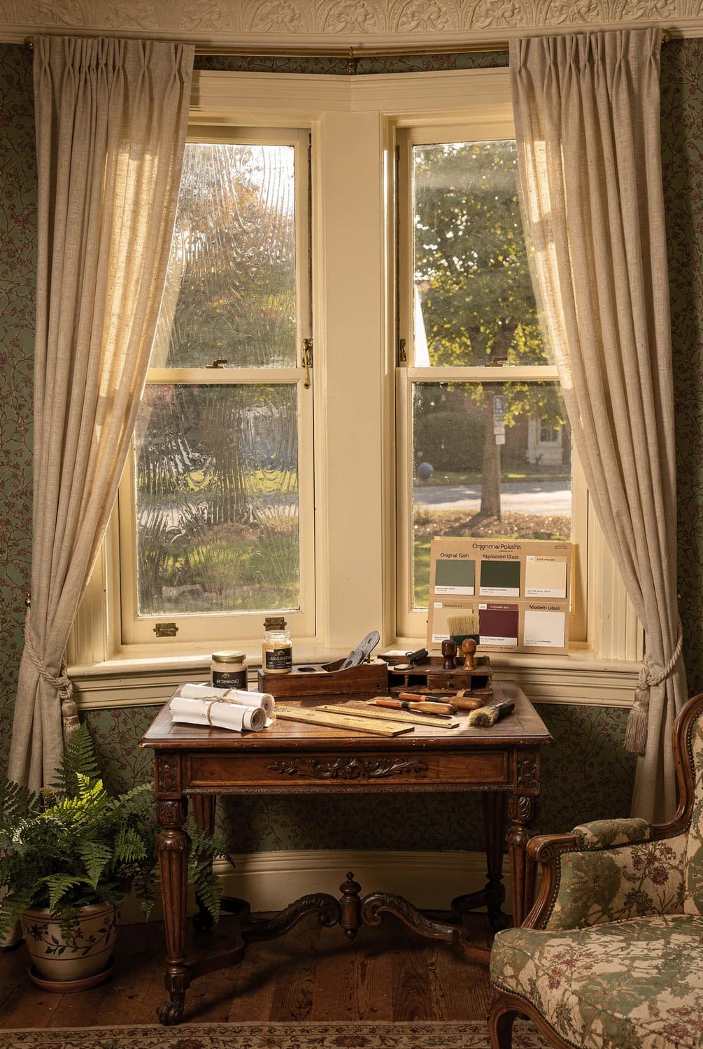 A restored Victorian bay window with textured glass, cream-painted wood frames, curtains, and a table displaying window restoration tools and finish samples.