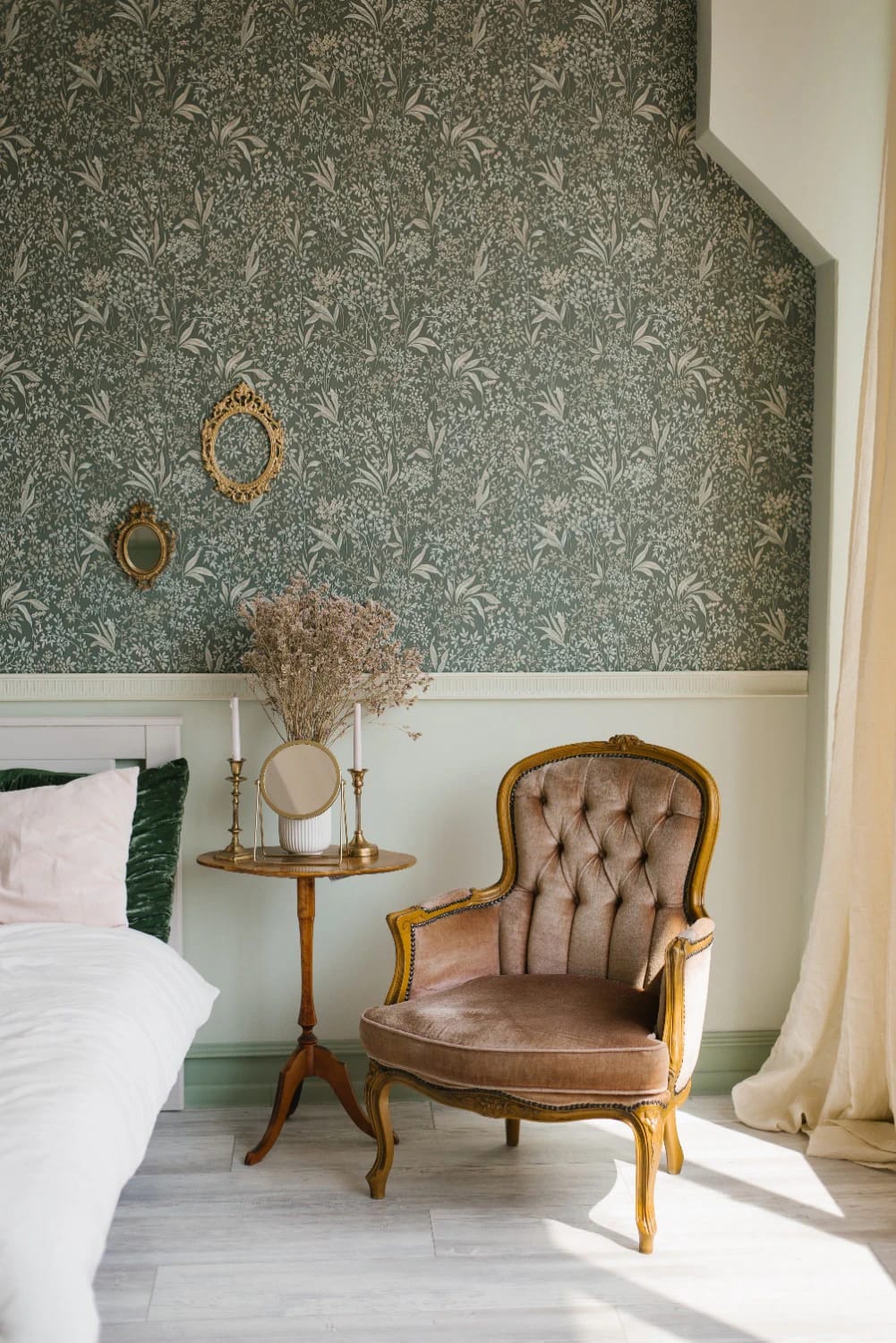 Victorian house interior featuring dark botanical wallpaper, two ornate oval mirrors, a tufted upholstered armchair, a small wooden table with candles and dried flowers, and part of a bed
