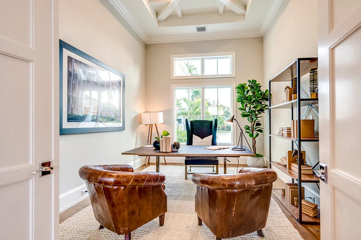 Warm home office with wood desk, stacked windows, leather chairs, open shelving, and decorative beamed ceiling