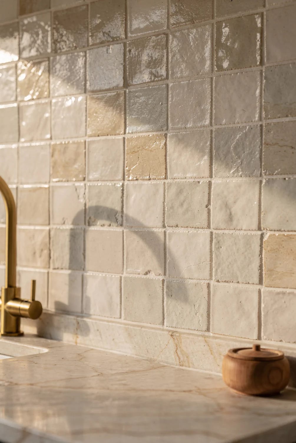 Warm Off-White Kitchen Backsplash Texture Close-up of a warm off-white kitchen backsplash made of glossy, uneven square tiles above a pale stone countertop, with a brass faucet and small wooden container visible