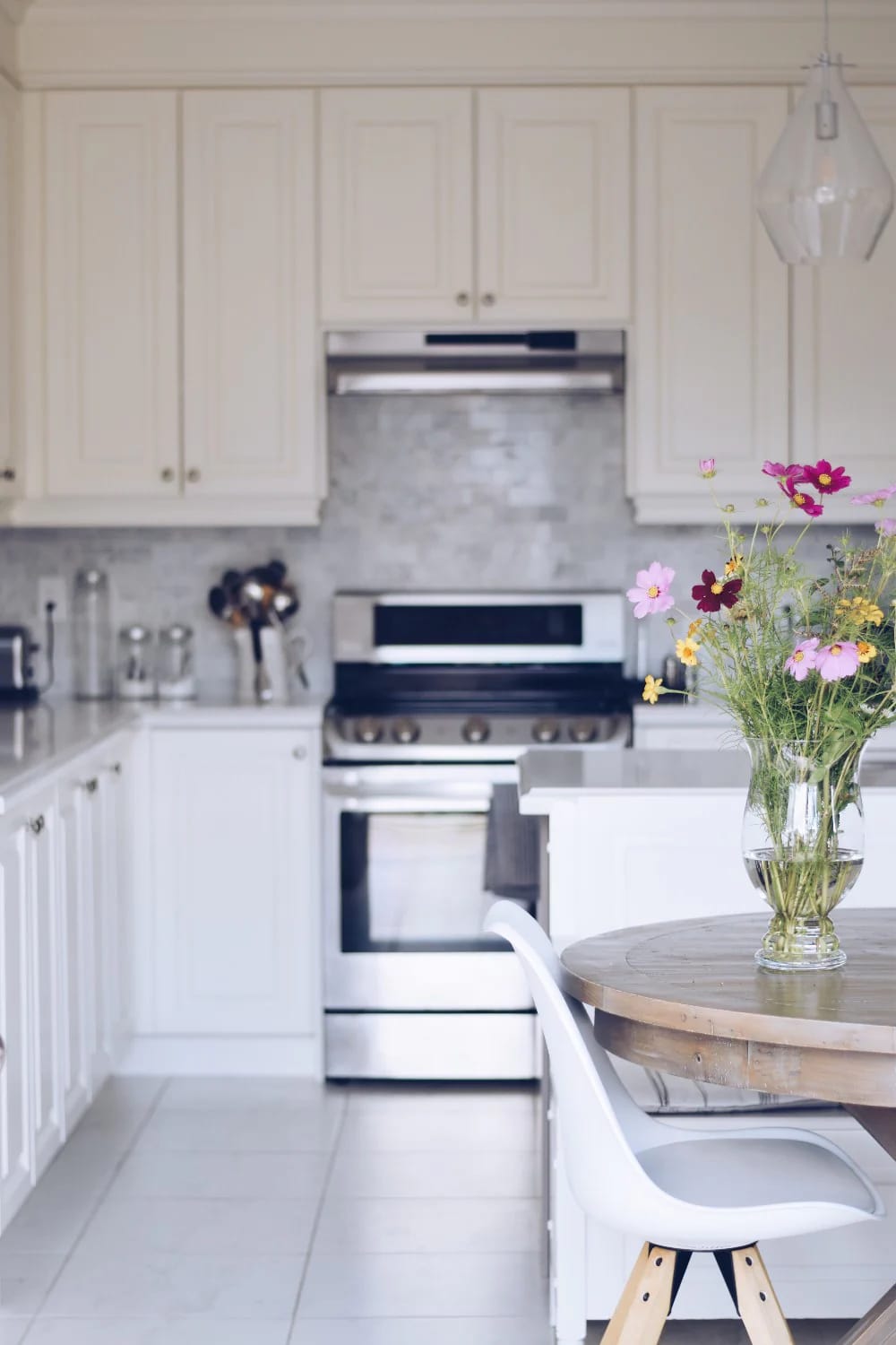 Warm Off-White Kitchen With Floral Dining Table Bright off-white kitchen with paneled cabinets, stainless steel range, pale backsplash, white island, and a round wooden table holding a vase of colorful flowers