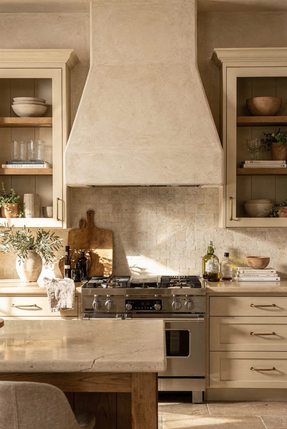 Warm Off-White Kitchen With Plastered Range Hood Warm off-white kitchen with a plastered range hood matching the surrounding cabinetry, open shelves, tiled backsplash, stainless steel range, and stone island.