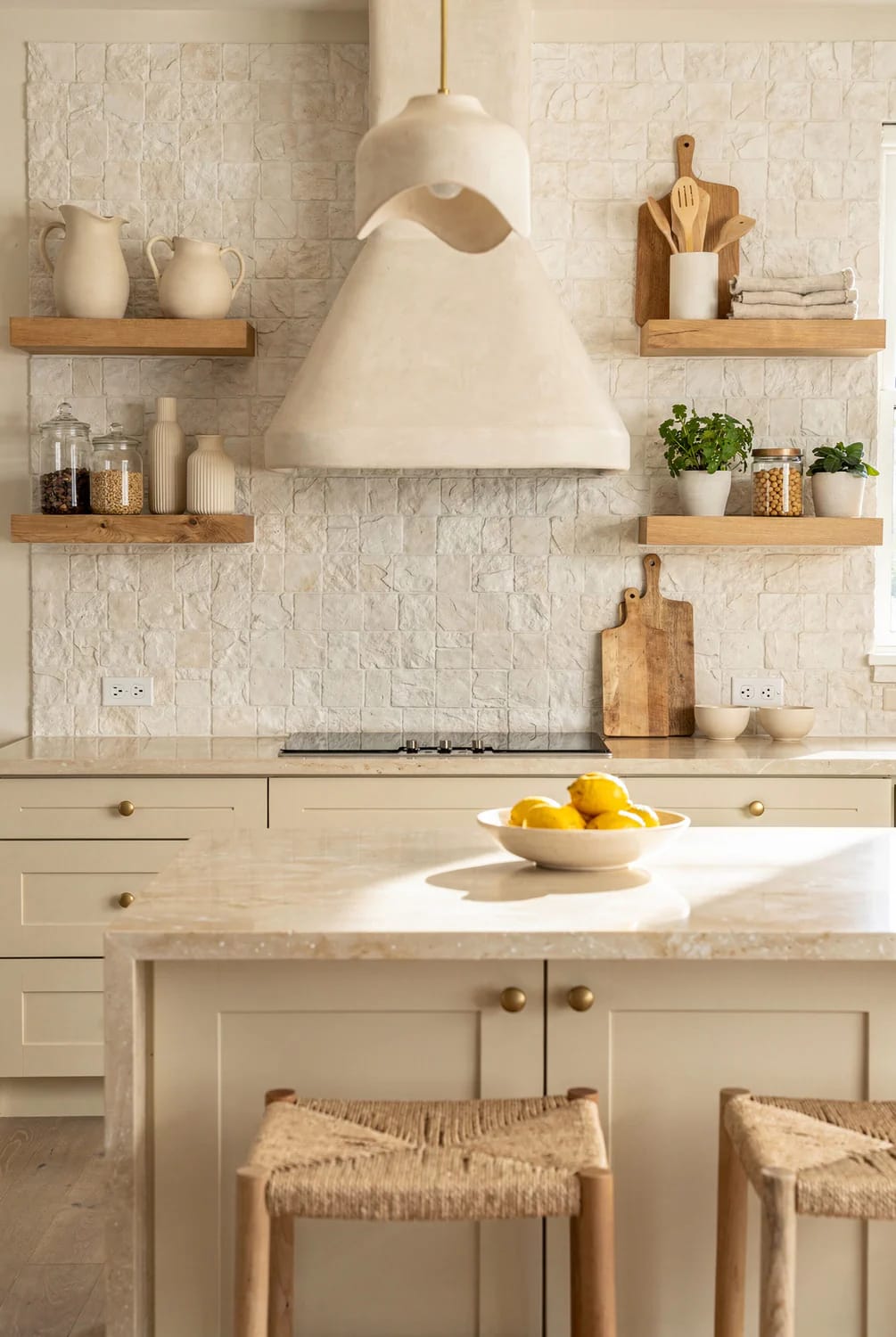 Warm Off-White Kitchen With Textured Backsplash Warm off-white kitchen featuring a textured light backsplash, wooden floating shelves, cream cabinetry, a large plaster range hood, and a bowl of lemons on the island