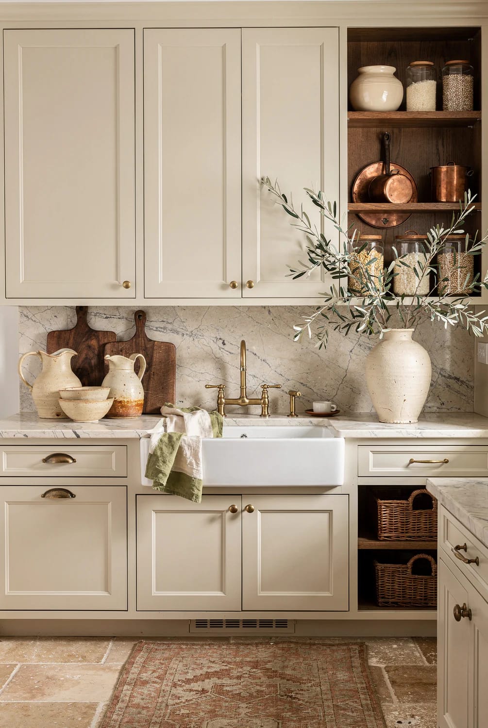 Warm Off-White Kitchen With Unlacquered Brass Hardware Warm off-white kitchen with paneled cabinets, unlacquered brass knobs and drawer pulls, a farmhouse sink, marble backsplash and countertop, open shelves with copper cookware and jars, and an olive branch in a ceramic vase