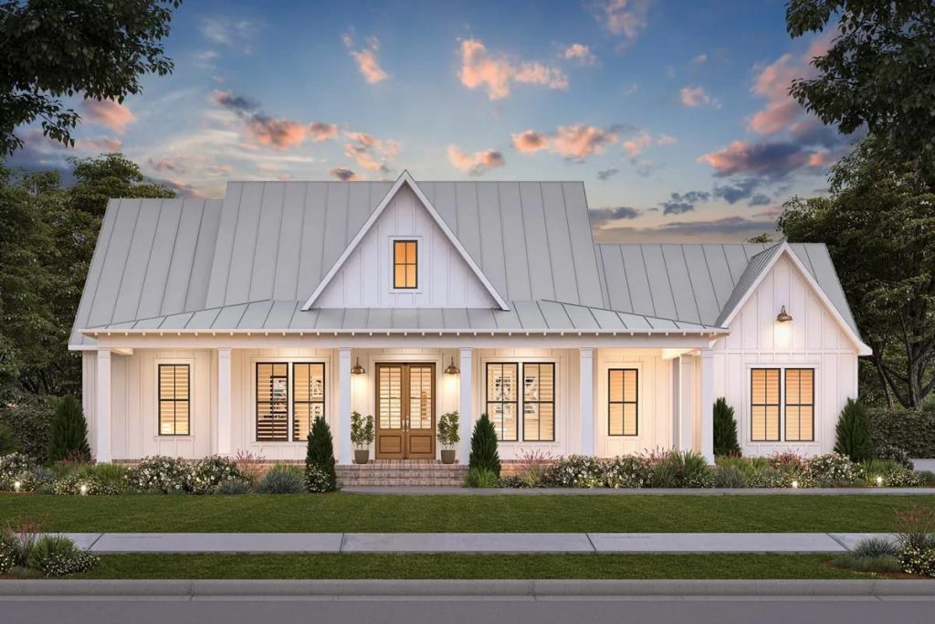 White cottage-style home with metal roof, centered porch entry, warm windows, and landscaped front garden at dusk