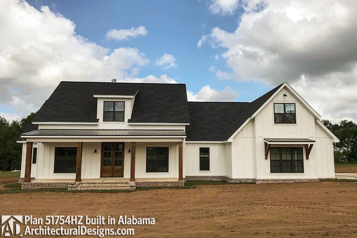 White farmhouse with covered porch, multiple gables, dark roof, wood columns, and brick foundation accents White farmhouse with covered porch, multiple gables, dark roof, wood columns, and brick foundation accents