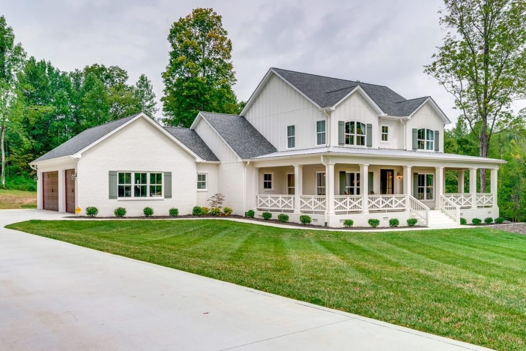 White farmhouse with gabled rooflines, covered porch, attached garage, lawn, and mature trees