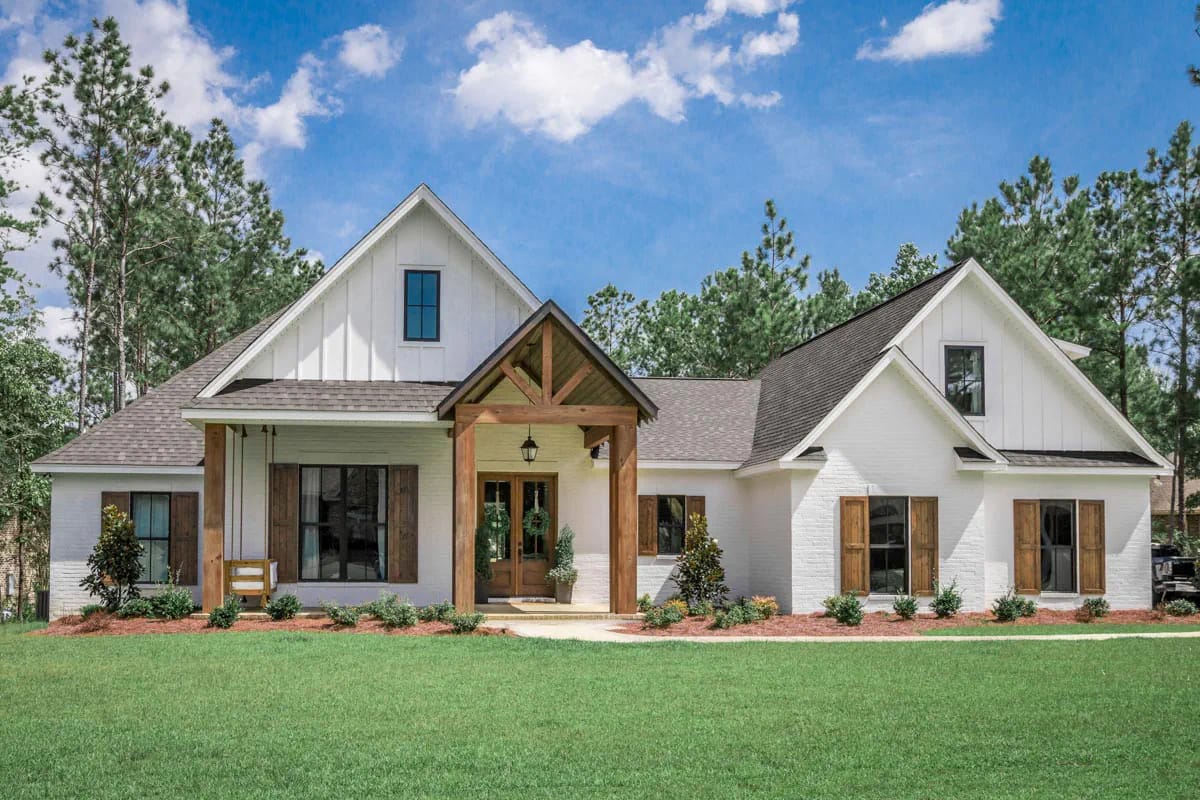 White gabled farmhouse with timber entry porch, dark windows, wood shutters, lawn, and pine trees