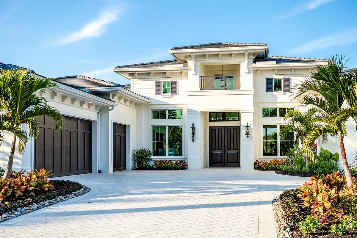 White stucco home with central balcony, dark doors, tiled roofs, tropical landscaping, and a broad paver drive