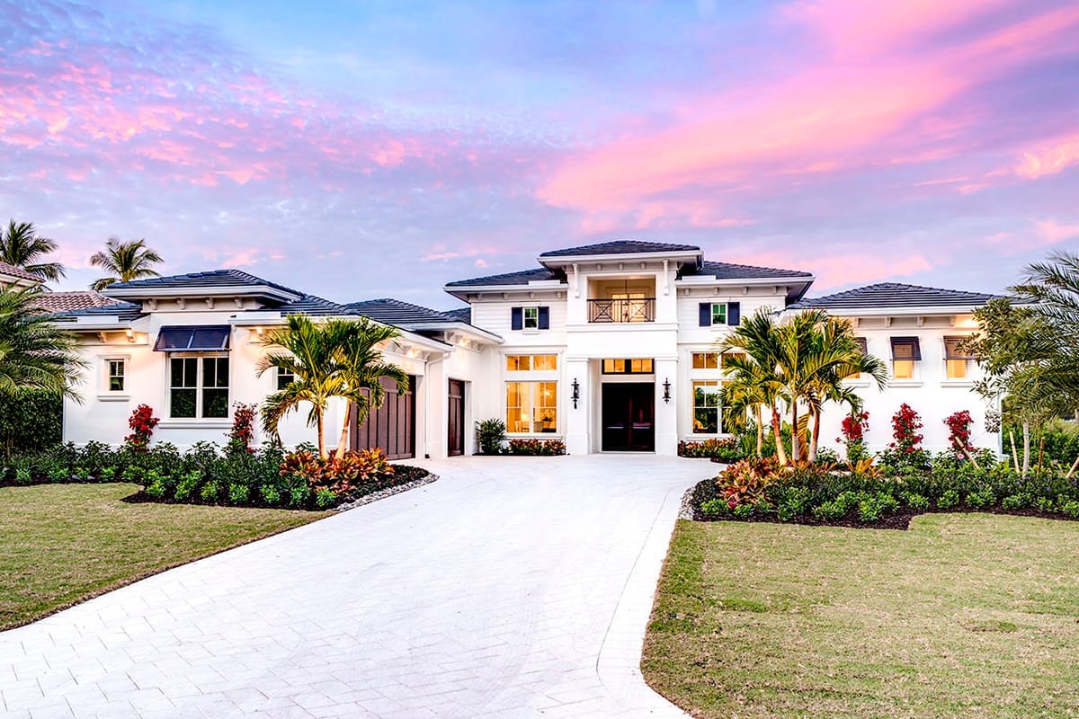 White stucco tropical-style home with dark tiled roofs, central balcony, palms, and a wide driveway at sunset