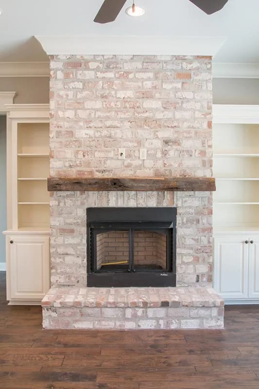 Whitewashed brick fireplace with dark wood mantel, built-in shelving, cabinets, and raised hearth