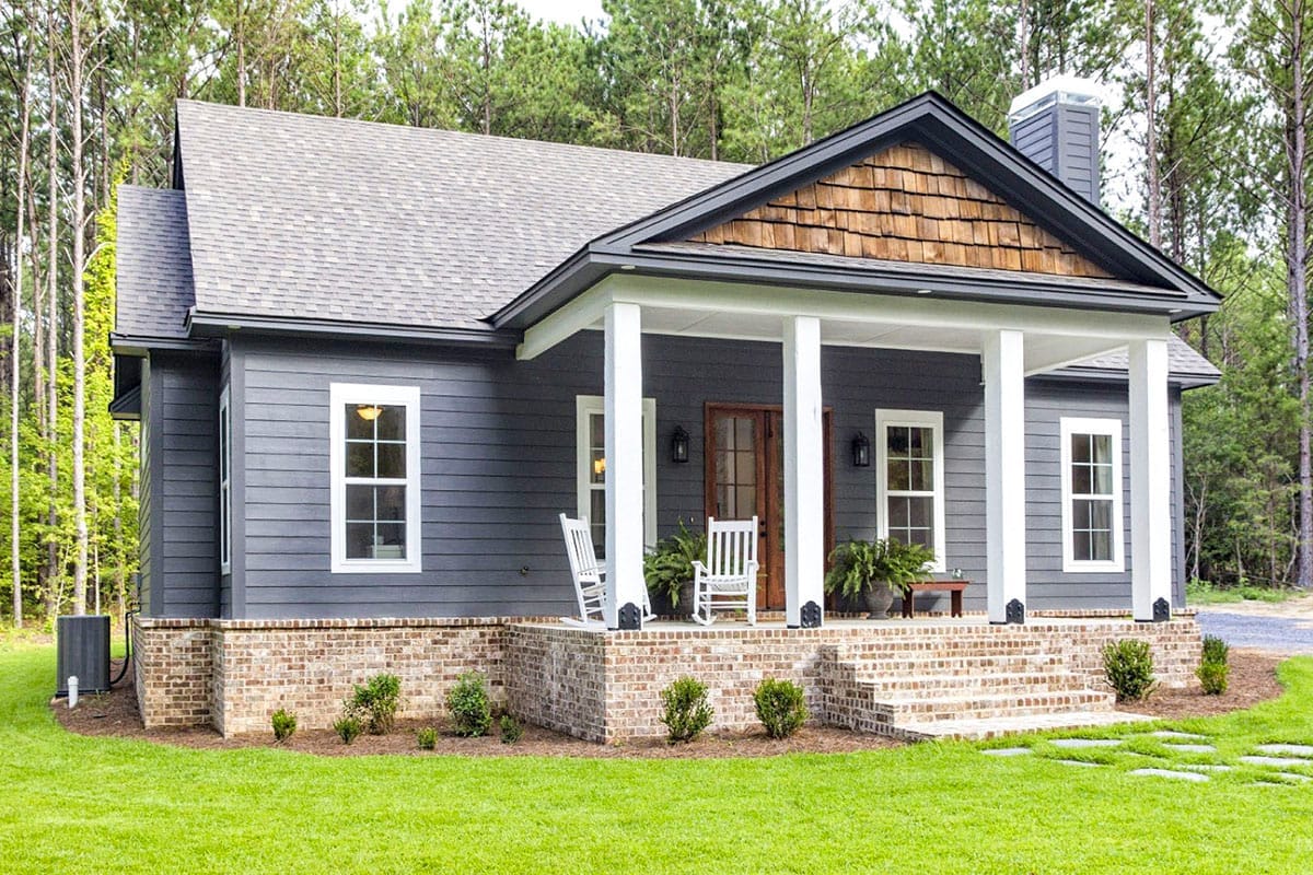 Dark gray cottage with a covered porch, brick steps, wood gable accent, and rocking chairs in a wooded setting Dark gray cottage with a covered porch, brick steps, wood gable accent, and rocking chairs in a wooded setting