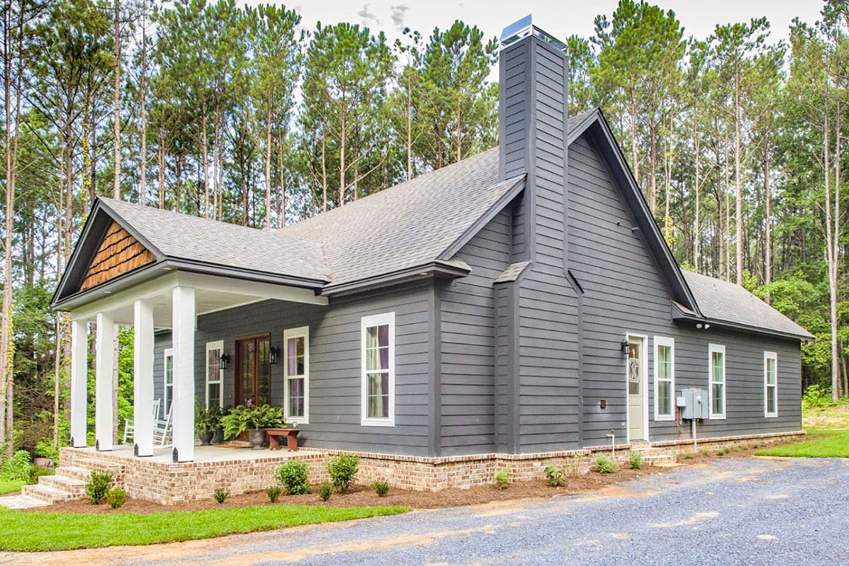 Gray cottage-style home with a covered porch, tall chimney, brick foundation, and dark pine-wood setting Gray cottage-style home with a covered porch, tall chimney, brick foundation, and dark pine-wood setting