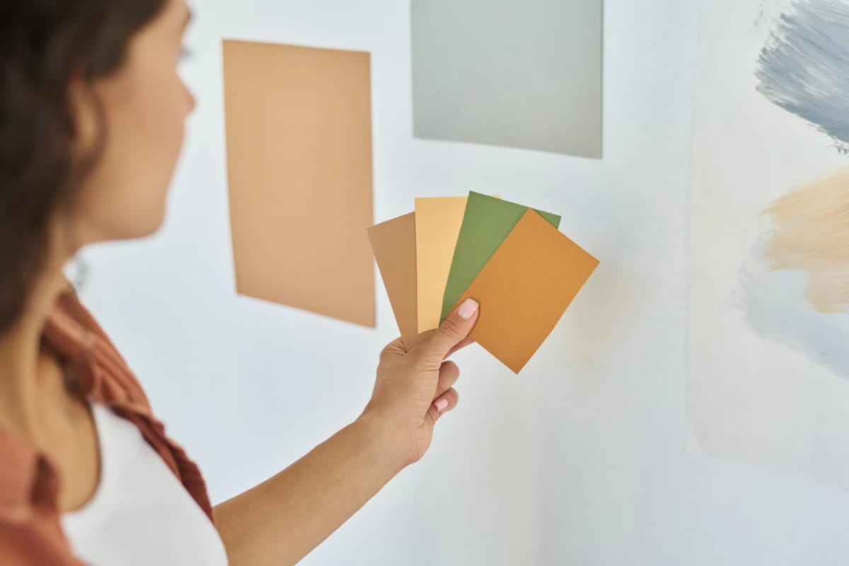 How To Test Paint Colors Before Buying Person holds several paint color cards near a wall with large beige, gray, green, and blue paint samples, illustrating how to test paint colors the right way before you buy