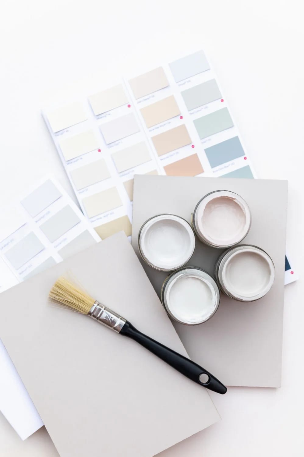 LRV Explained With Paint Chips And Supplies Paint color swatches, four open cans of light-colored paint, blank boards, and a paintbrush arranged for an LRV paint color comparison
