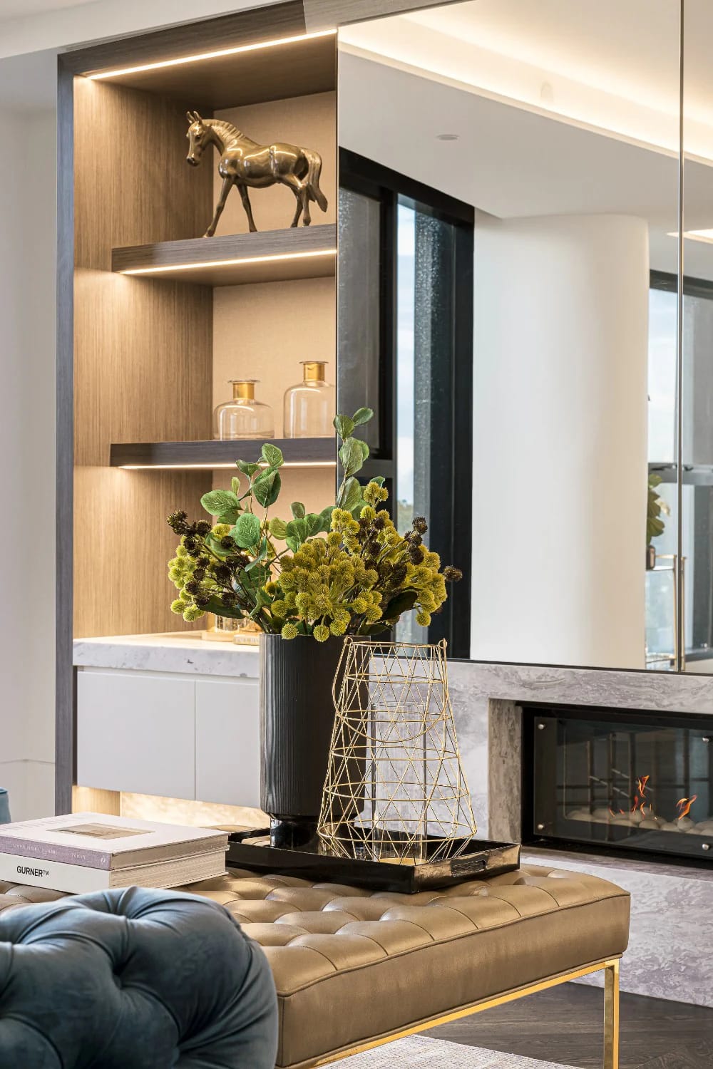 Elegant living room featuring illuminated wood display shelves with a bronze horse sculpture and glass bottles, a large mirror, fireplace, floral arrangement, tufted ottoman, and decorative tray.