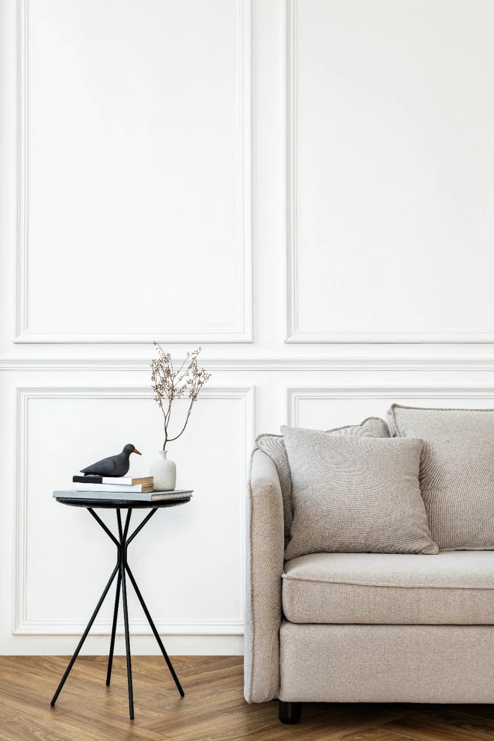 A beige sofa with textured cushions sits beside a slim black side table holding stacked books, a dark bird sculpture, and a white vase with dried branches. White paneled wall molding and herringbone wood flooring create a refined living room setting for rich living room ideas.