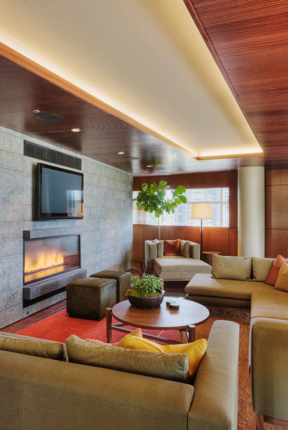 Rich living room featuring beige sofas and chairs around a round wooden coffee table, a gray stone wall with fireplace and television, warm wood paneling, recessed lighting, a potted plant, and a red rug