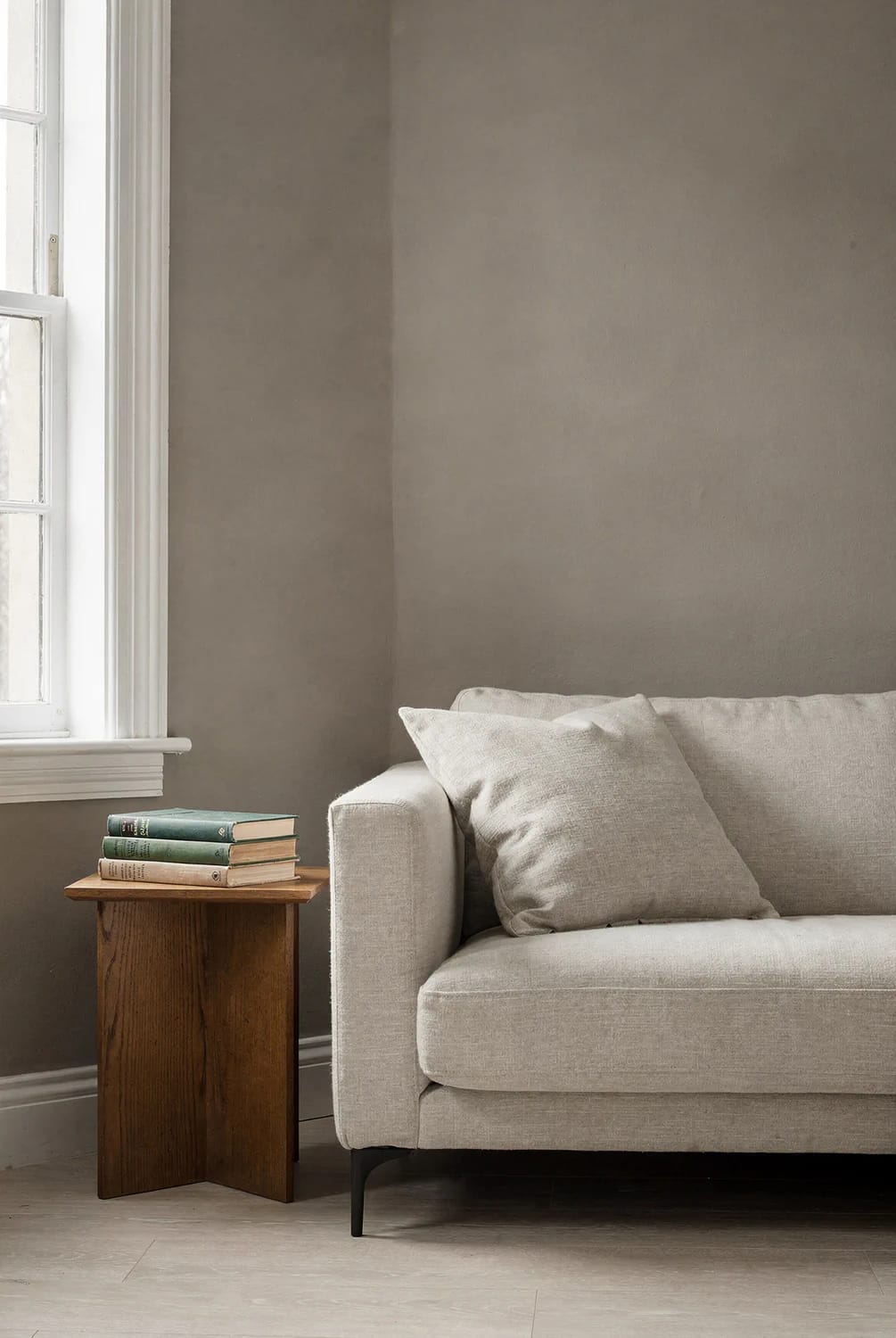 Room Orientation And Paint Color Lighting A gray-painted living room wall beside a white-trimmed window, with natural light falling over a beige sofa, wooden side table, and stacked books, illustrating how room orientation affects paint colors