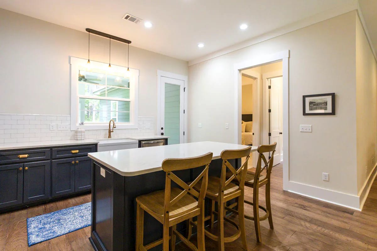 Storybook Bungalow Kitchen Island Seating Three Kitchen in the Storybook Bungalow house plan with a large island, three wooden bar stools, dark blue cabinets, white subway tile backsplash, farmhouse sink, and pendant lights