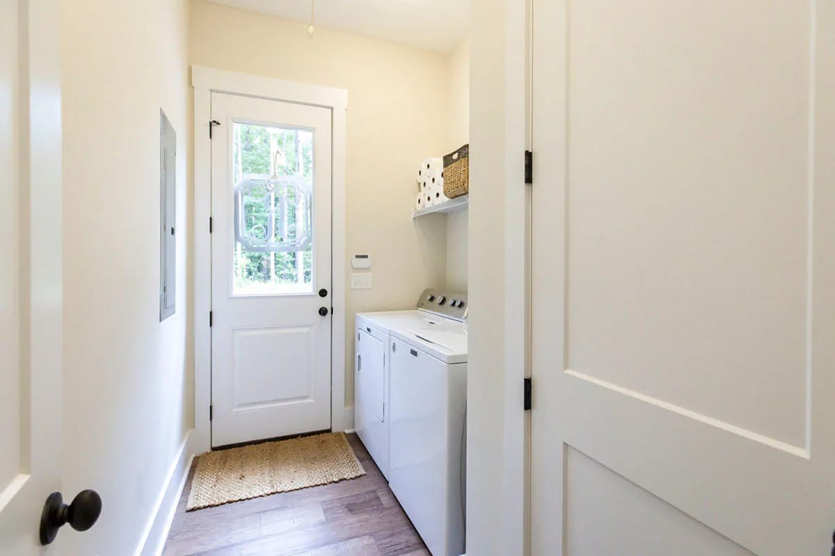Storybook Bungalow Laundry Room Side Yard Door Laundry room in the Storybook Bungalow house plan with a white washer and dryer, shelving, a woven mat, and a glass-paneled door opening to the side yard