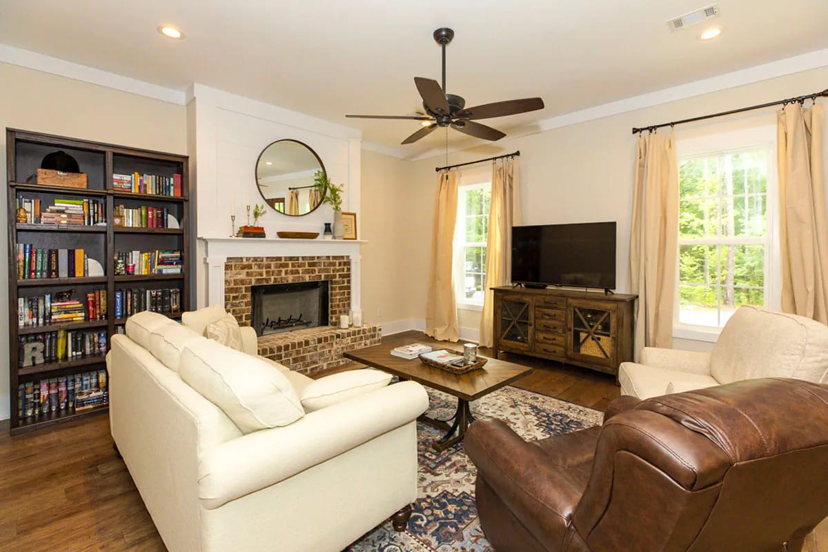 Storybook Bungalow Living Room With Fireplace Storybook bungalow living room with a brick fireplace, built-in bookshelves, cream sofas, a leather chair, television, coffee table, ceiling fan, and windows with curtains