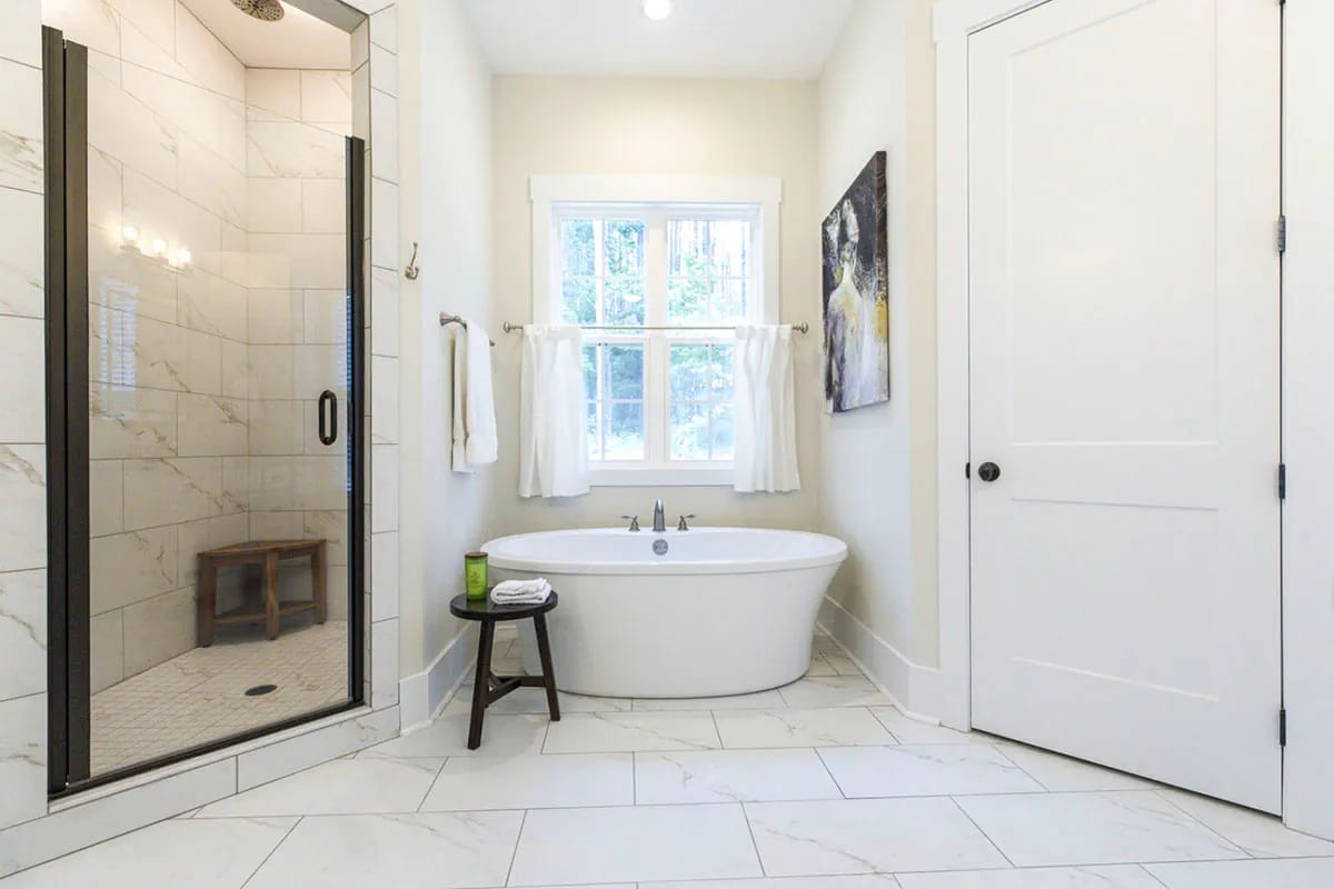 Storybook Bungalow Master Bathroom Design Bright master bathroom in the Storybook Bungalow house plan featuring a freestanding white bathtub beneath a window, a glass-enclosed tiled shower, white marble-look floor tiles, and a closed white door