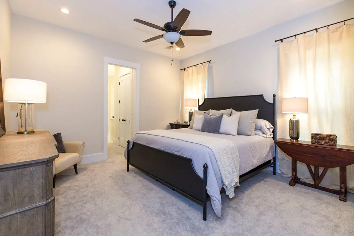 Storybook Bungalow Master Bedroom Master bedroom in the Storybook bungalow house plan with a black four-poster bed, light bedding, bedside tables, lamps, curtained windows, ceiling fan, desk, and carpeted floor