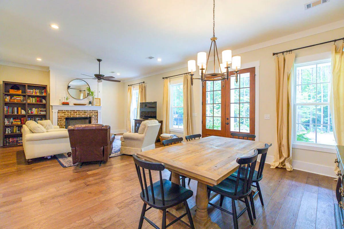 Storybook Bungalow Open Living And Dining Room Open-plan living and dining area in the Storybook Bungalow, featuring a brick fireplace, built-in bookcase, seating, large wood dining table, chandelier, and windows with French doors