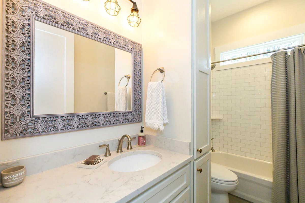 Storybook Bungalow Shared Bathroom Design Shared bathroom in the Storybook Bungalow house plan with a marble-look vanity, ornate framed mirror, white sink, toilet, and tiled bathtub with a shower curtain