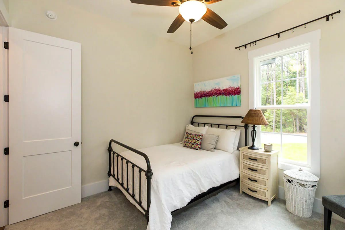 Storybook Bungalow Square Bedroom Square bedroom in the Storybook Bungalow house plan with a black metal bed, white bedding, bedside chest, window, ceiling fan, artwork, and laundry basket