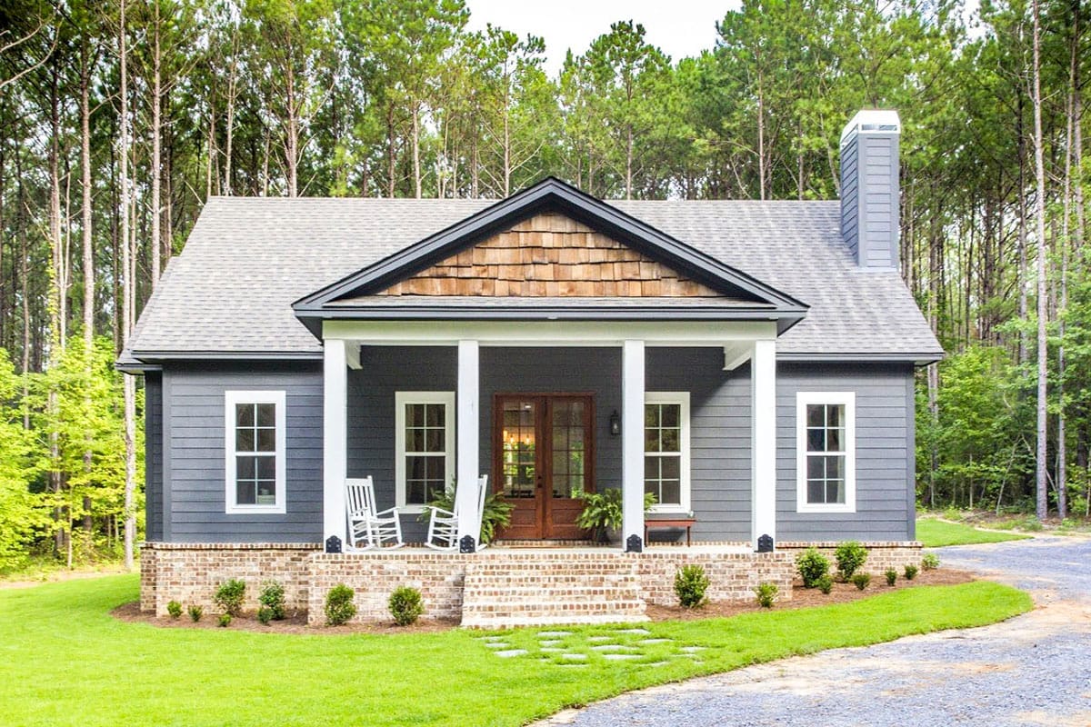 Storybook Bungalow 3-Bedroom House Plan with Large Front and Back Porches (1,631 Sq. Ft. Floor Plan) 1 Symmetrical gray cottage with a white-columned porch, brick foundation, wood-shingle gable, and pine forest setting